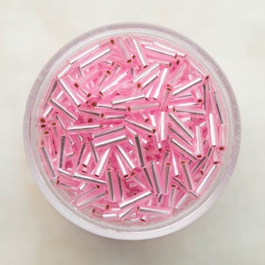 Pode incluir: Um recipiente de vidro transparente cheio de contas de vidro rosa e prateadas. As contas são pequenas e de forma cilíndrica.