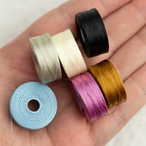 Peut inclure: Six bobines de fil de diff&eacute;rentes couleurs, dont bleu clair, rose clair, beige clair, blanc, or et noir. Les bobines sont dispos&eacute;es dans une main.