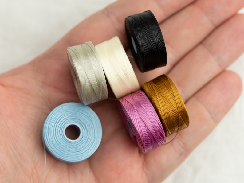 Peut inclure: Six bobines de fil de diff&eacute;rentes couleurs, dont bleu clair, rose clair, beige clair, blanc, or et noir. Les bobines sont dispos&eacute;es dans une main.
