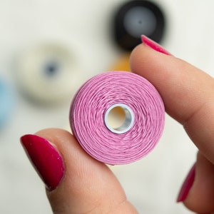Peut inclure: Gros plan d'une bobine de fil rose tenue par une main avec du vernis &agrave; ongles rouge.