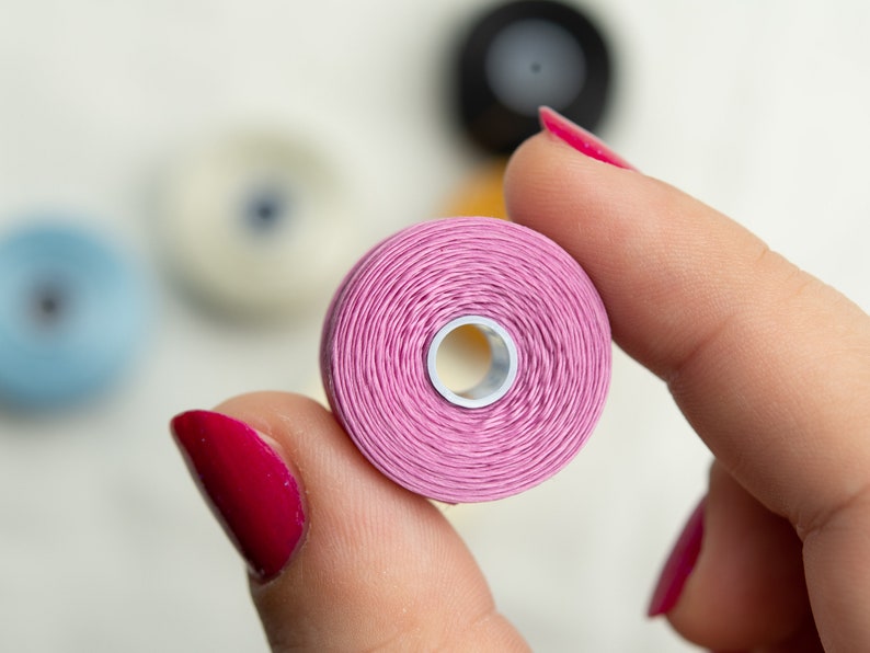 Peut inclure: Gros plan d'une bobine de fil rose tenue par une main avec du vernis &agrave; ongles rouge.