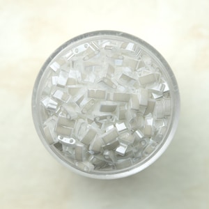 May include: A clear glass jar filled with small, silver-colored metal beads. The beads are rectangular in shape and have a slightly rounded edge.
