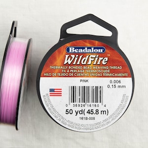 Może przedstawiać: Szpula różowego Beadalon WildFire, termicznie łączonego, nici do tkactwa koralikowego. Etykieta podaje, że ma grubość 0,006 cala (0,15 mm) i zawiera 50 jardów (45,8 metrów) nici. Etykieta informuje również, że jest przeznaczona wyłącznie do użytku przez osoby dorosłe.