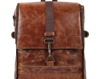 May include: A brown leather backpack featuring a fold-over flap closure secured with a dark brown fabric strap and a metal hook clasp. The bag includes a top handle, side metal D-rings, and visible stitching details across the front panel.