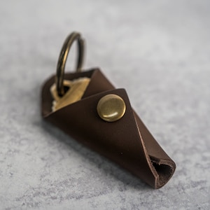 Könnte beinhalten: Ein brauner Leder-Schlüsselanhänger mit einem Messing-Druckknopfverschluss und einem Messing-Schlüsselring. Der Schlüsselanhänger ist in der Mitte gefaltet und der Schlüsselring ist sichtbar.