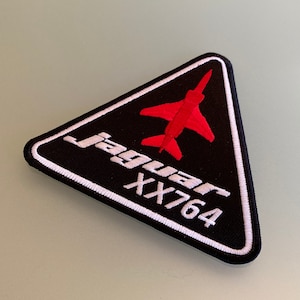 May include: A black triangular patch with white stitching. The patch features a red silhouette of a jet aircraft and the text "Jaguar XX764" in white.