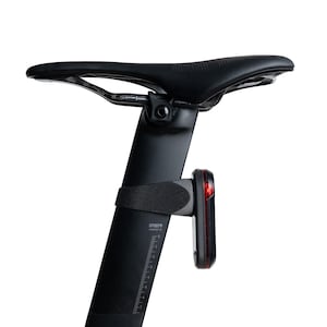 May include: A black bicycle seat mounted on a vertical seat post. The seat post features a printed scale with markings in centimeters and the text SP0079. A small red LED safety light is attached to the post with a black rubber strap.