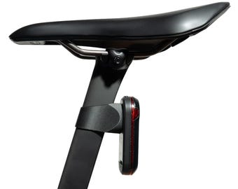 Könnte beinhalten: Ein schwarzer Fahrradsattel auf einer Sattelstütze mit einem vertikalen schwarzen Rücklicht, das am Rohr befestigt ist. Der Sattel hat eine glatte, matte Oberfläche und ist über ein Metallschienensystem mit einer sichtbaren Einstellschraube verbunden.