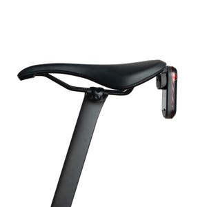Könnte beinhalten: Ein schwarzer Fahrradsattel auf einer schwarzen Sattelstütze mit schlankem, aerodynamischem Profil und strukturierter Oberfläche. Ein kleines, rechteckiges, schwarz-rotes Rücklicht ist sicher an den Sattelstreben befestigt.