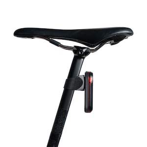 May include: A black bicycle seat mounted on a black seat post with a vertical rectangular red LED safety light attached to the post via a black rubber strap. The light features a thin red border and is positioned directly below the saddle.