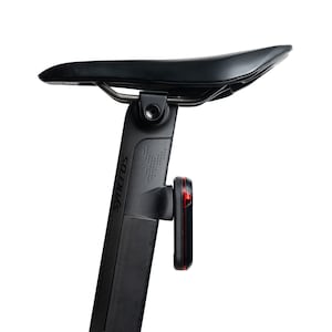 May include: A black bicycle seat mounted on a black aerodynamic seatpost with the brand name Syncros printed vertically in black. A small rectangular black rear light with a red lens is attached to the back of the seatpost structure.
