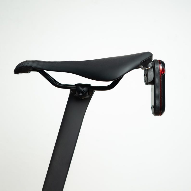 May include: A black bicycle saddle mounted on a black seat post with a vertical red rear light attached to the back of the seat. The saddle features a textured surface and is supported by black metal rails connected to the seat post clamp.