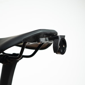 May include: A black bicycle saddle featuring a mounted black plastic accessory bracket attached to the rear rails. The bracket has a circular interface with a central locking mechanism designed for securing cycling equipment to the seat.