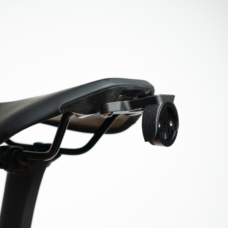 May include: A black bicycle saddle featuring a mounted black plastic accessory bracket attached to the rear rails. The bracket has a circular interface with a central locking mechanism designed for securing cycling equipment to the seat.