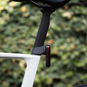 May include: Close-up of a bicycle seat and frame. The black seat is mounted on a black seat post. A red and black taillight is attached to the seat post. The frame is white.