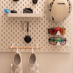 Pode incluir: Um organizador de parede de painel perfurado branco com uma prateleira contendo um ciclocomputador preto, uma luz vermelha e uma câmara de ar. Um capacete MET branco, dois pares de óculos de ciclismo coloridos, chaves, um estojo e sapatilhas.