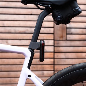 Könnte beinhalten: Nahaufnahme eines weißen Rennrads mit schwarzem Sattel, Sattelstütze und Rücklicht. Eine schwarze Satteltasche ist unter dem Sattel befestigt. Das Fahrrad hat einen schwarzen Reifen und einen Holzuntergrund.