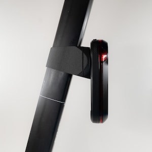 May include: A black bicycle tail light with a red light is mounted on a black metal frame. The light is attached to the frame with a black bracket. The background is plain white.