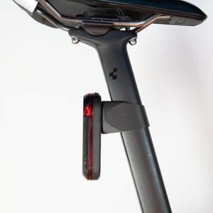 Puede incluir: Una luz trasera Garmin negra y roja fijada a una tija de sillín negra. La luz trasera tiene una luz roja y está montada con un soporte negro. El sillín de la bicicleta es negro con raíles plateados.