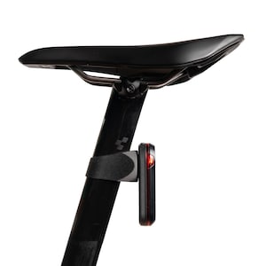 以下が含まれることがあります： 黒い自転車用サドルと黒いシートポスト。シートポストには黒いブラケットを介して小型の長方形の赤いLED安全ライトが取り付けられています。シートポストの上部付近には小さな灰色のロゴがあり、ライトは点灯しています。