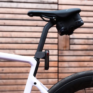 May include: A white bicycle frame with a black seat post, a black saddle, and a black under-seat storage bag. A rectangular black rear light with a red lens is mounted vertically on the seat post below the saddle, positioned above the rear wheel.