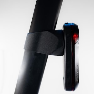 Könnte beinhalten: Ein schwarzes Fahrradlicht ist mit einem schwarzen Riemen an einem schwarzen Fahrradrahmen befestigt. Das Licht hat eine rote Linse und einen schwarzen Körper. Das Licht ist mit einem schwarzen Riemen am Rahmen befestigt.