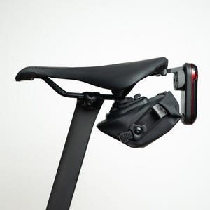 May include: A black bicycle saddle mounted on a seat post with a black under-seat storage bag attached. A red rear bicycle light is secured to the back of the saddle rails, positioned vertically behind the storage bag on a mounting bracket.