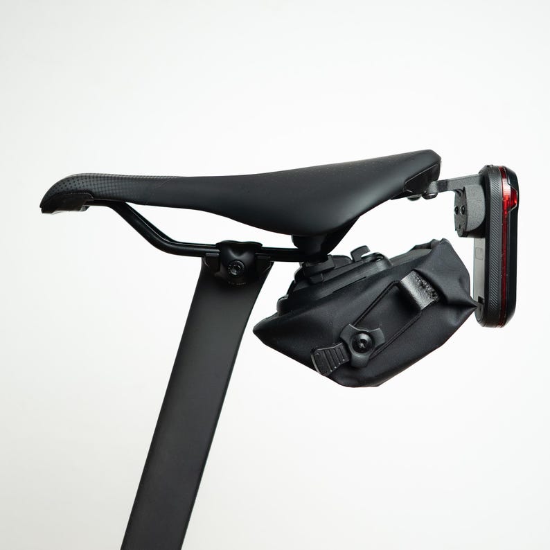 May include: A black bicycle saddle mounted on a seat post with a black under-seat storage bag attached. A red rear bicycle light is secured to the back of the saddle rails, positioned vertically behind the storage bag on a mounting bracket.