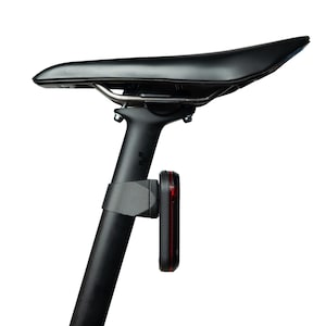 Pode incluir: Um selim de bicicleta preto montado num espigão de selim preto, com um design elegante e aerodinâmico. Fixada ao espigão está uma luz de segurança traseira retangular vermelha com moldura preta, presa por um suporte de montagem integrado.