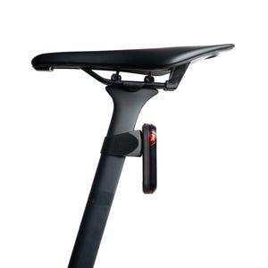 May include: A black bicycle saddle mounted on a black seat post with a small rectangular red LED safety light attached to the post via a black strap. The light is positioned vertically, showing a thin red light strip on its outer edge.