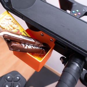 Op de afbeelding: Een oranje plastic snackhouder bevestigd aan een zwarte hometrainerconsole. De houder bevat twee chocoladerepen en een gele candybar. Het stuur en de console van de fiets zijn zichtbaar.