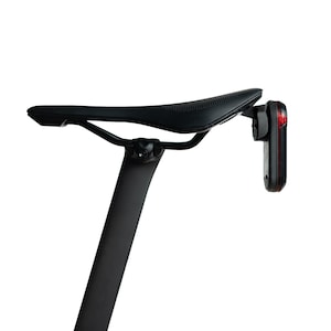 May include: A black bicycle saddle mounted on a vertical seat post, featuring a slim, aerodynamic profile with a textured surface. A rectangular red LED safety light is attached to the rear of the saddle, positioned vertically for visibility.