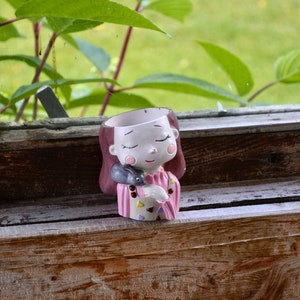May include: A ceramic planter shaped like a girl with closed eyes, holding a small gray rabbit in her arms. She is wearing a pink dress with colorful accents.