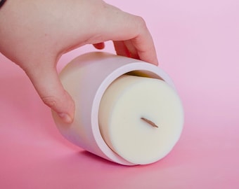 May include: A white candle with a wooden wick is inside a white ceramic container. The container is being held by a hand.