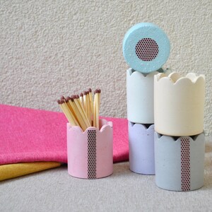以下が含まれることがあります： 波打つ縁のある小さなコンクリート製の装飾容器が5つあります。容器は、薄い青、白、ラベンダー、ピンクなど、さまざまなパステルカラーでペイントされています。各容器には、細く黒と茶色の模様のストライプがあります。1つの容器には、木製のマッチの山が入っています。
