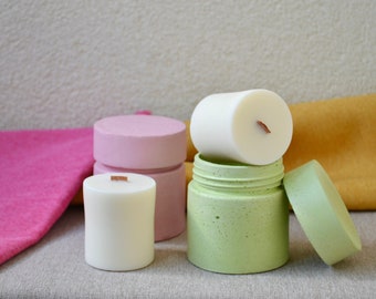 May include: A collection of handmade candles in various colors. The candles are white and cylindrical, with wooden wicks. They are placed on and next to a green container and a pink container. A pink and yellow fabric are in the background.