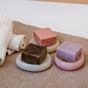 May include: Four bars of soap on small round ceramic dishes. The soaps are in shades of pink, brown, and purple. The dishes are in shades of gray and pink. The soaps are on a brown fabric surface.