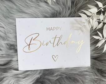 May include: A white greeting card with the words "HAPPY Birthday" in gold script, along with a small heart. The card has a dotted pattern and is set against a gray, fluffy background with white floral accents.