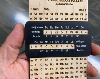 May include: A wooden musical cypher tool called the Harmonator, with a black and white chart showing musical scales, chords, and solfege. The tool is made of wood and has a black and white chart with the text "Harmonator" and "olddognewtricks.club".