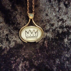 May include: A gold necklace with a round pendant featuring a black crown design. The pendant is set in a clear glass dome.