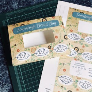 May include: A Sourdough Bread Bag sewing pattern printed on paper with a beige background, floral motifs, and blue text. The design features a rectangular cutout, line art of bread loaves, and care instructions. The paper measures up to 19 inches.