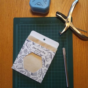 Puede incluir: Una bolsa de pan de lino blanca con una ilustración en blanco y negro de pan en la parte delantera. La bolsa está sobre una estera de corte verde con una regla y un perforador.