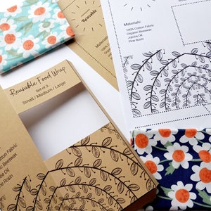 Puede incluir: Un juego de tres envoltorios de alimentos reutilizables en tamaños pequeño, mediano y grande. Los envoltorios están empaquetados en una caja de cartón marrón con un diseño floral. Se ven dos envoltorios de tela con estampados de margaritas en azul y turquesa.