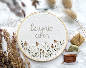 May include: A white embroidery hoop with the name "Laynie Ann" embroidered in brown thread. The hoop is surrounded by a border of green and brown embroidered flowers.