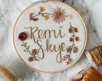 May include: Embroidered hoop art with the names "Remi Skye" in gold thread surrounded by pink and orange flowers.
