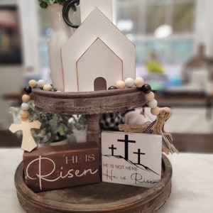 Op de afbeelding: Een houten dienblad met twee niveaus met een witte houten kerk, een houten kruis en twee houten borden met de tekst "Hij is opgestaan" en "Hij is niet hier, Hij is opgestaan". Het dienblad is versierd met een touwtje met bruine en witte kralen.