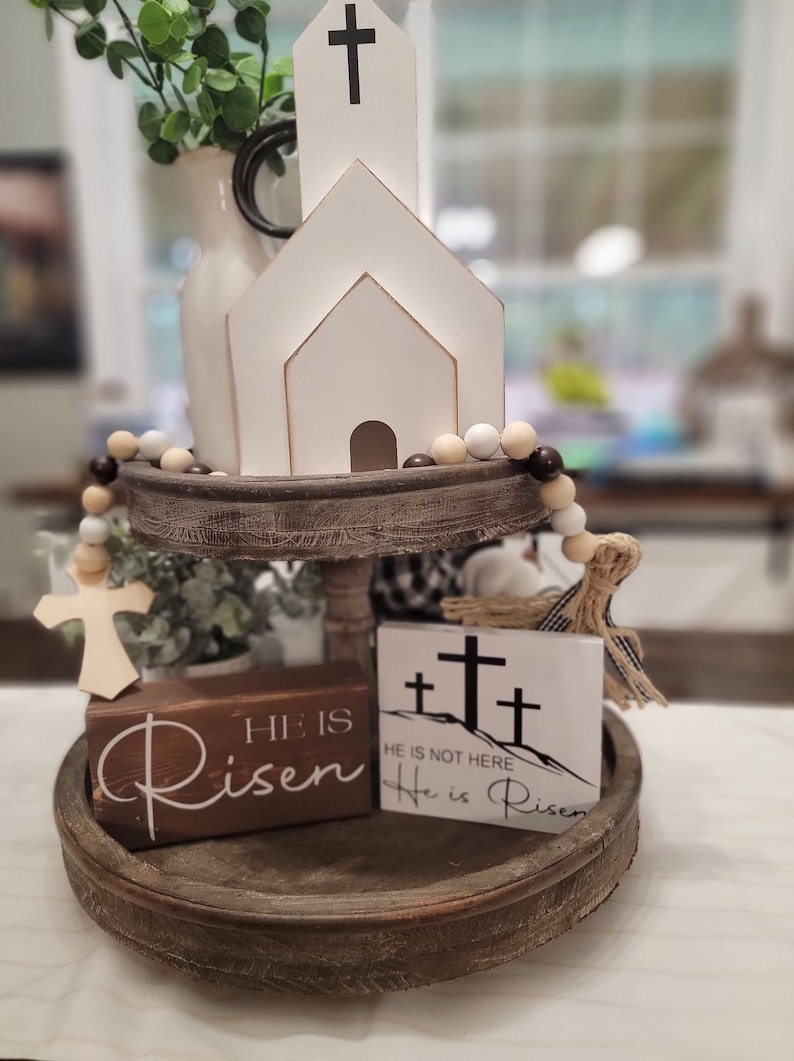 Op de afbeelding: Een houten dienblad met twee niveaus met een witte houten kerk, een houten kruis en twee houten borden met de tekst "Hij is opgestaan" en "Hij is niet hier, Hij is opgestaan". Het dienblad is versierd met een touwtje met bruine en witte kralen.