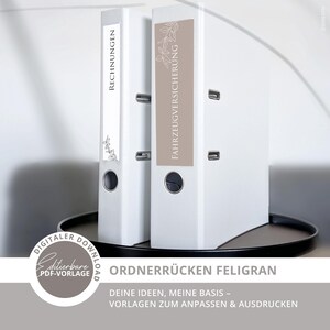 May include: Two white ring binders with labels. One label reads "RECHNUNGEN" and the other reads "FAHRZEUGVERSICHERUNG". Both labels have a floral design. The binders are on a dark tray. Text at the bottom reads "ORDNERRÜCKEN FELIGRAN".