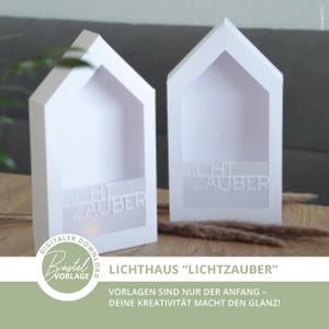 Puede incluir: Dos cajas decorativas blancas en forma de casa con el texto "LICHT ZAUBER" en la parte delantera. Las cajas están hechas de un material blanco mate y tienen un recorte en el centro. Las cajas están sobre una superficie de madera.