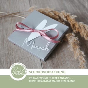 May include: A gray gift box tied with a pink ribbon and a white floral accent. The word "Mensch" is written across the box in a cursive font. The box is on a wooden surface, with dried plants in the background. The image also contains the text "SCHOKOVERPACKUNG".